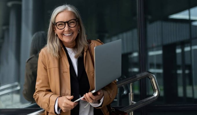 Smiling businesswoman holding a laptop outside an office building, ready to work or attend a meeting.